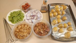 Marinated beef and chicken cubes with chopped bell peppers, red onion, cherry tomatoes, and parboiled potatoes prepared for grilled kabobs on a charcoal kettle grill