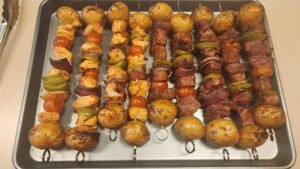 Grilled beef and chicken kabobs with potatoes, bell peppers, red onion, and cherry tomatoes resting after cooking on a charcoal kettle grill