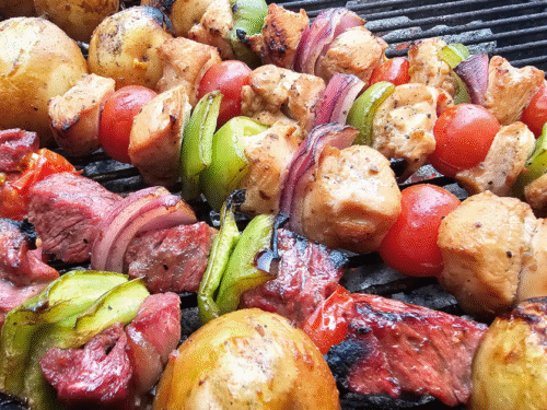 Grilled beef and chicken kabobs with bell peppers, red onion, and potatoes cooked over charcoal on a kettle grill