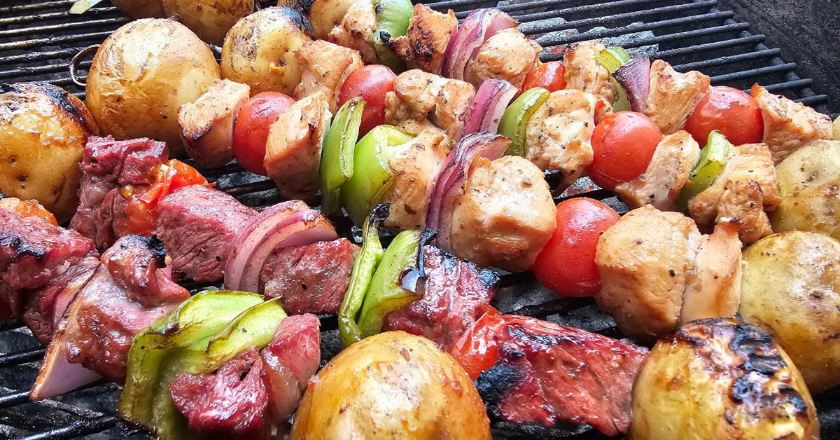 Grilled beef and chicken kabobs with bell peppers, red onion, and potatoes cooked over charcoal on a kettle grill