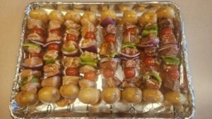 Assembled beef and chicken kabobs with potatoes, bell peppers, red onion, and cherry tomatoes arranged on skewers before grilling on a charcoal kettle grill