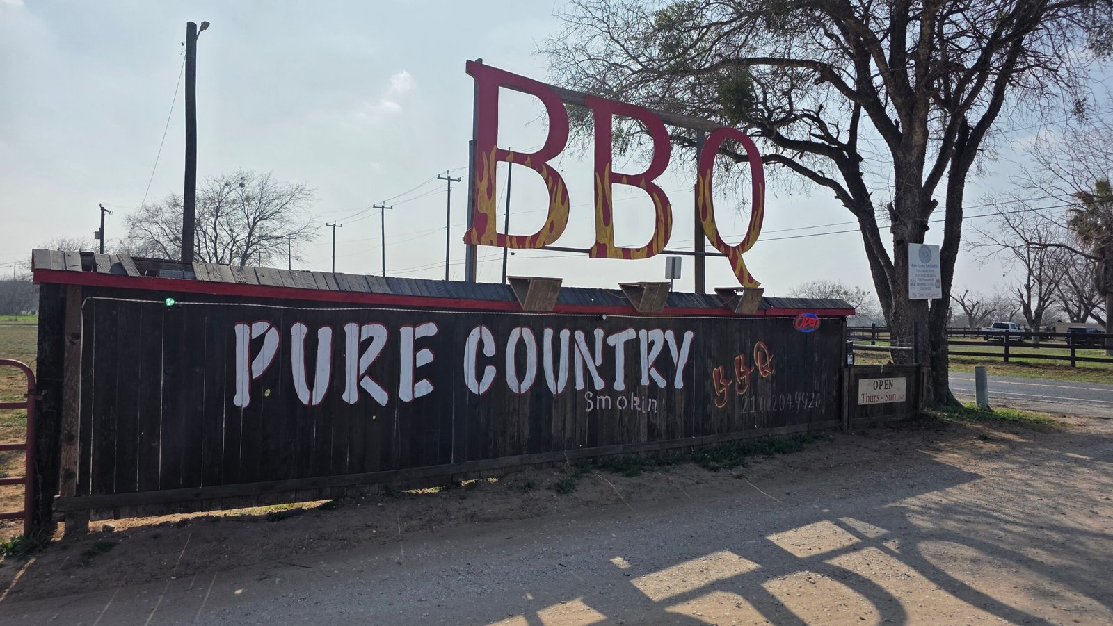 Pure Country BBQ roadside sign in Pleasanton Texas with large red BBQ letters and rustic wooden fence backdrop