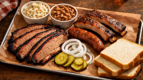 Balanced Texas BBQ tray with sliced brisket ribs sausage pickles onions white bread coleslaw and pinto beans on butcher paper